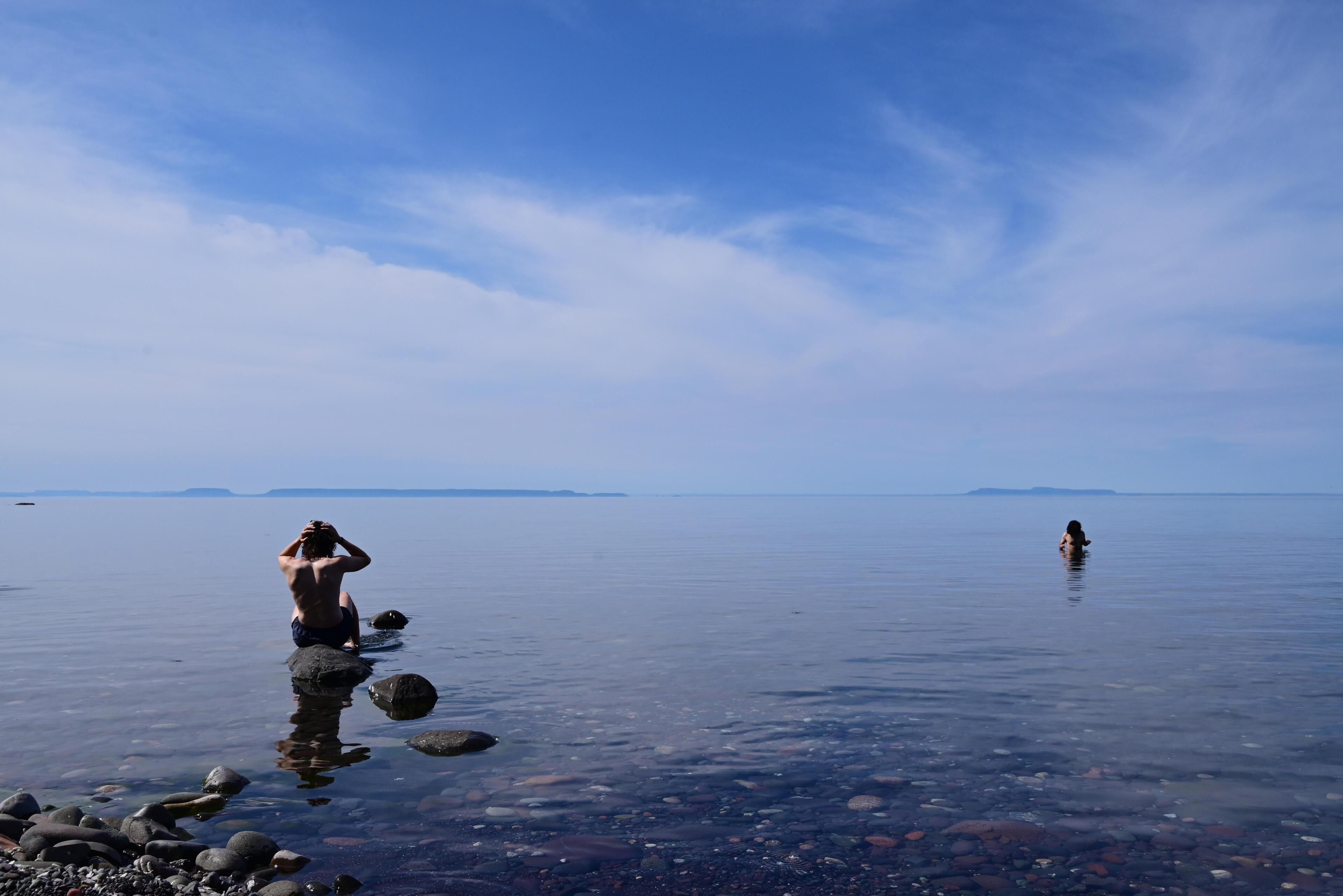 Two backpackers swim in calm, glass like, water in Lake Superior with only rocks in and under the water and the distant Canadian shore visible