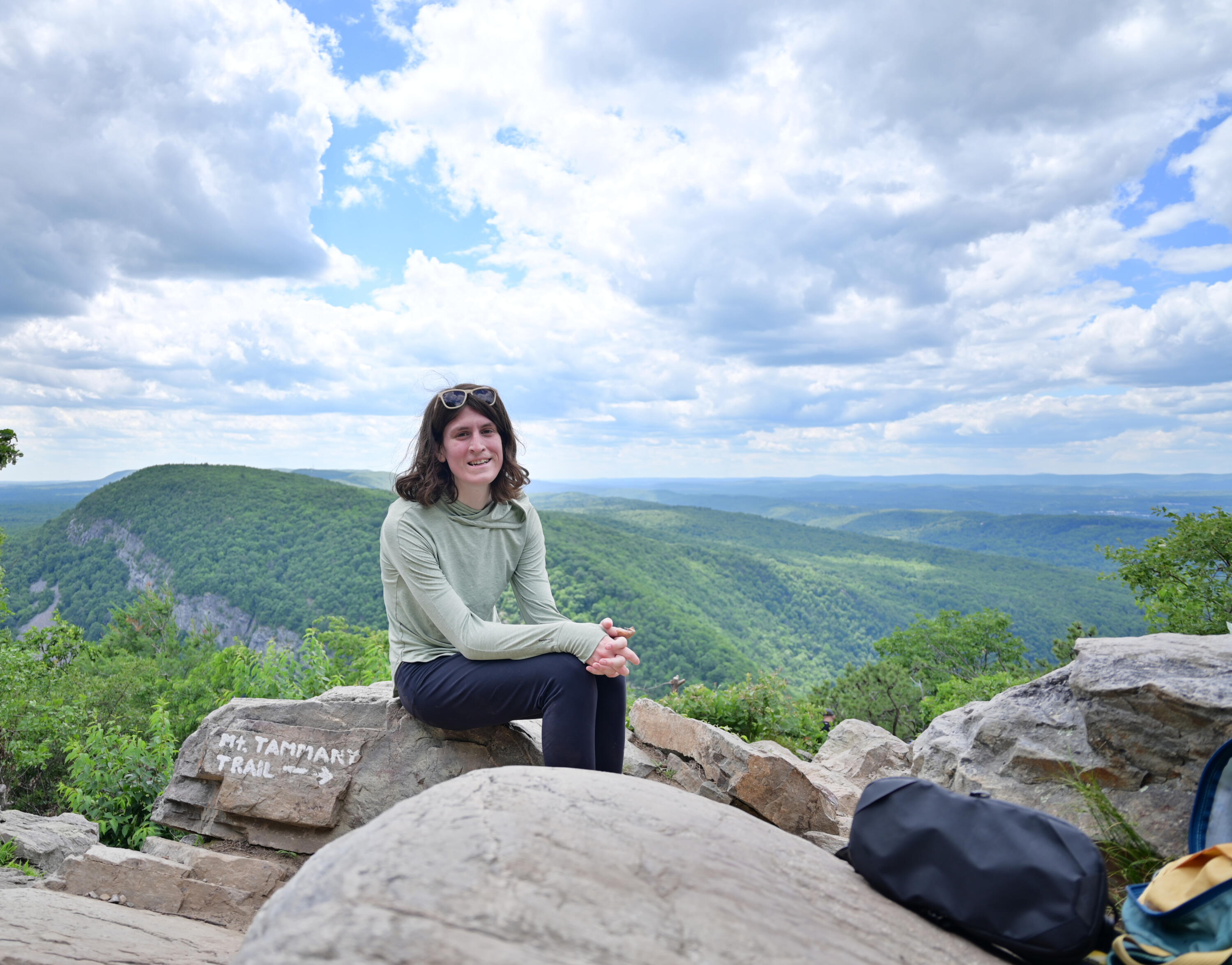 Your Guide on Mt Tammany A young person with shoulder length hair wearing a camo green sun hoodie and sunglasses in their hair sits on a rock with the words "Mt. Tammany Trail" painted on it. In the distance, another mountain, Mt Minsi, is visible and beyond that the rolling lands