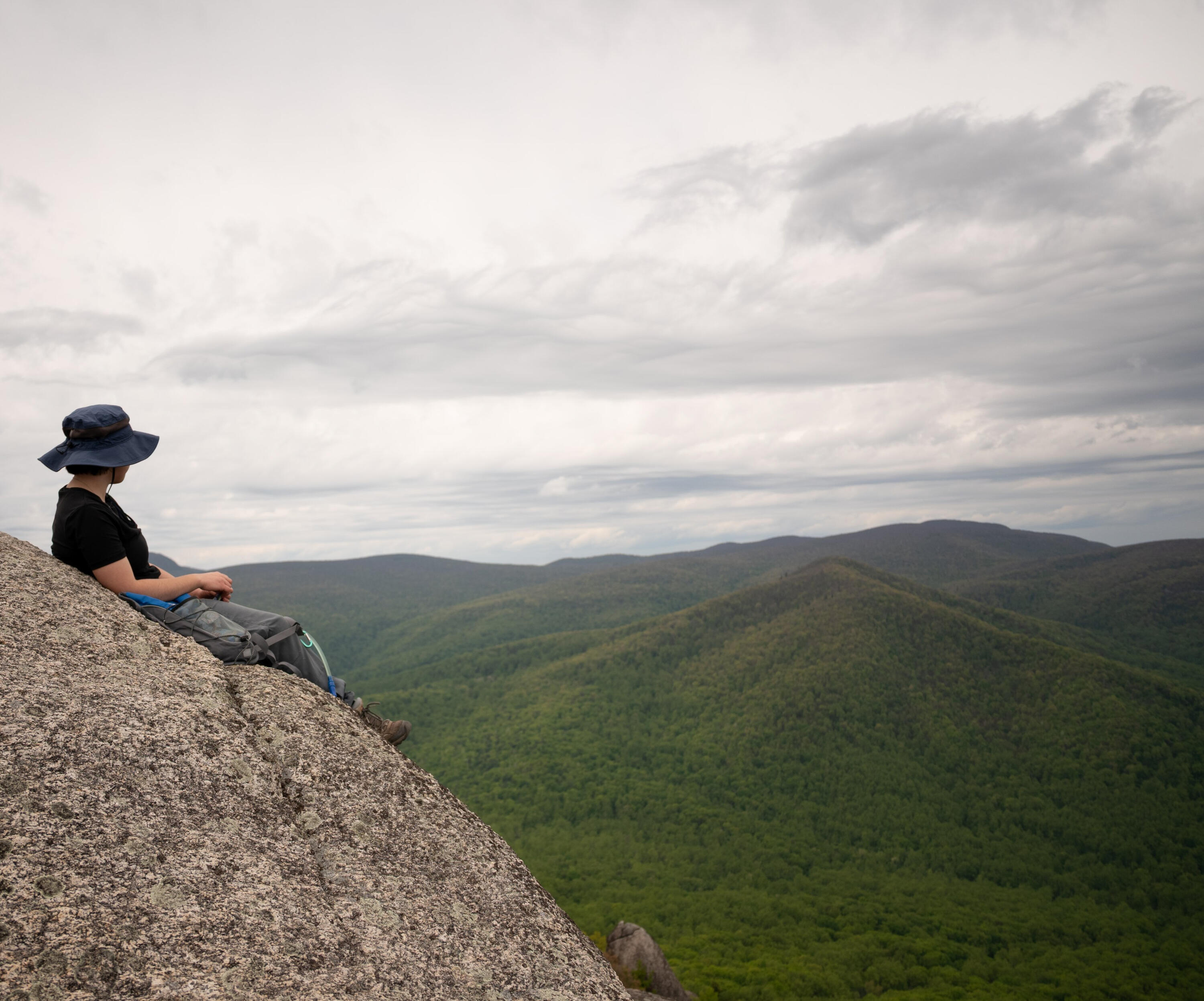 A hiker takes a break on top of a mountaintop boulder and looks out at the Shenandoah mountains